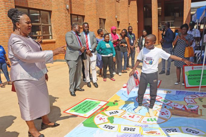 A child representative demonstrates to the Honourable Minister of Gender, Jean Muonaouza Sendeza, how game boards are used to raise awareness about climate change during her visit to the Save the Children pavilion at the National Children’s Summit on Climate Crisis and Child Rights.